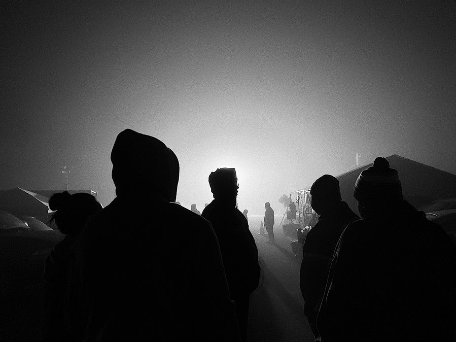 Silhouetted figures in hoods stand in fog before a bright light at night, in black and white