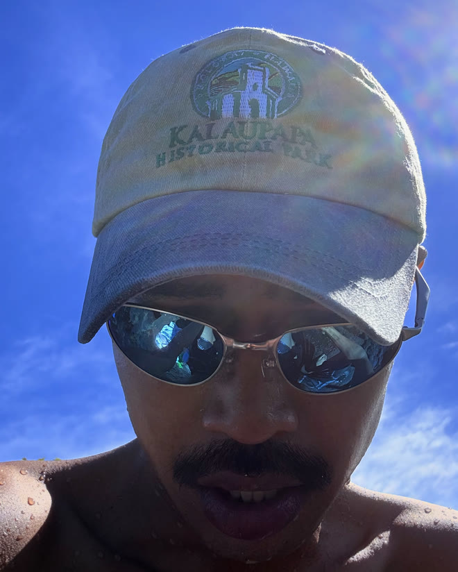 Sky Bruno in a Kalaupapa National Historical Park cap and mirrored sunglasses, against a blue sky